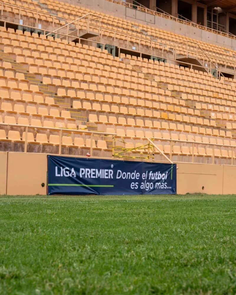 Tribuna del Estadio ITO con butacas amarillas y una lona de la Liga Premier junto a la cancha.