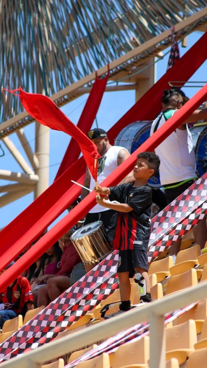 Un niño con la playera rojinegra de Dragones ondea una bandera roja en la tribuna, junto a la batucada.