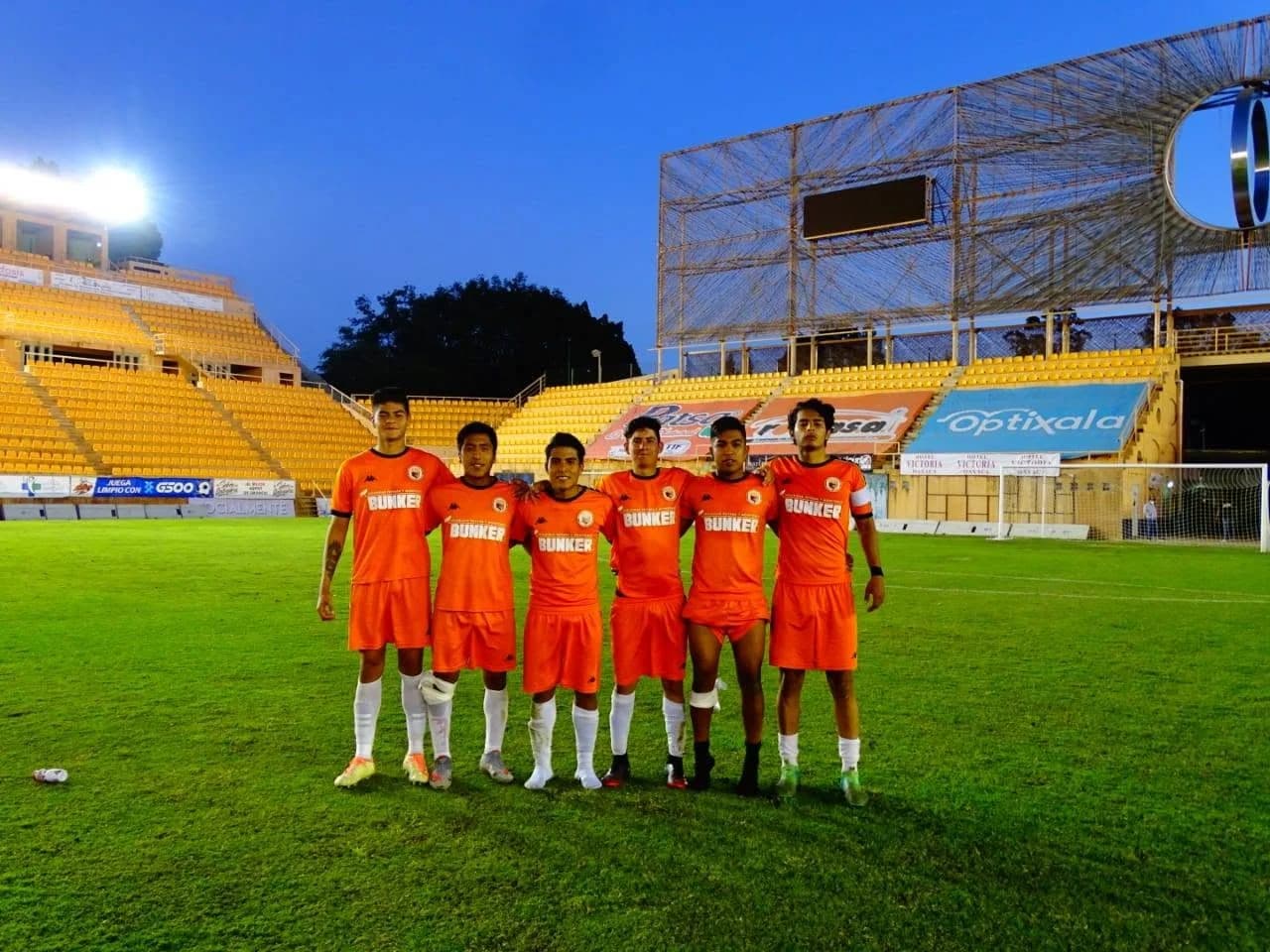 Seis jugadores de Dragones posan abrazados sobre el césped del estadio, al anochecer.