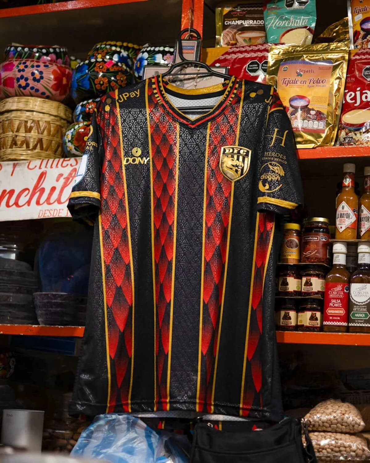 La camiseta rojinegra de local de Dragones, colgada en un puesto de mercado oaxaqueño entre barro, chocolate y salsas.