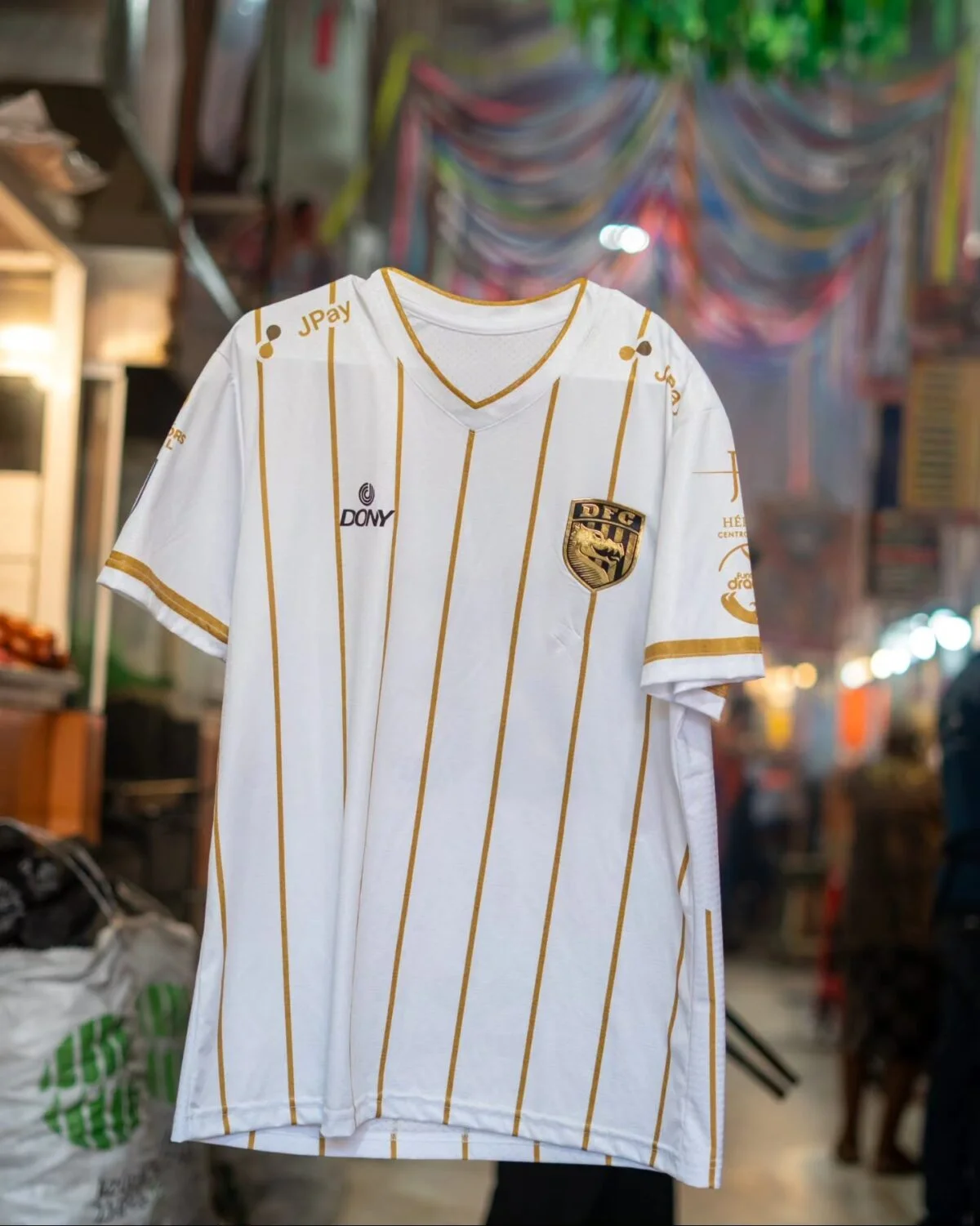 La camiseta blanca de visita de Dragones, con líneas doradas, colgada en un pasillo de mercado bajo papel picado.